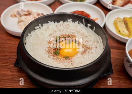 Authentischer koreanischer Rettichreis in einer heißen Steinschale mit rohem Eigelb und Sesamsamen, serviert mit traditionellen Beilagen auf einem hölzernen Esstisch. Stockfoto