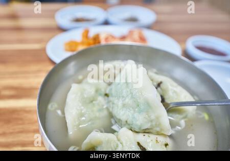 Authentische nordkoreanische übergroße Knödel in einer warmen, klaren Brühe, serviert in einem Metallbecken mit Algengarnitur auf einem hölzernen Restauranttisch. Stockfoto