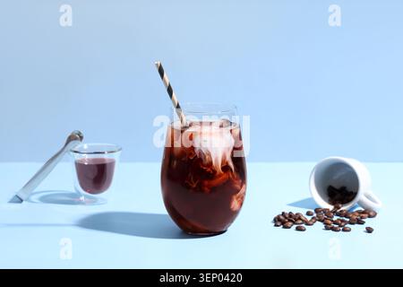 Erfrischender Eiskaffee mit Creme Swirl und Kaffeebohnen auf hellblauem Hintergrund, Eiskaffee mit cremigem Swirl und gestreiftem Strohhalm sitzt in einem cl Stockfoto