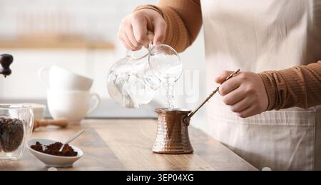 Person, die Wasser in Kupfercezve für die türkische Kaffeezubereitung auf hölzernem Arbeitsplatte mit Kaffeebohnen gießt, weiße Keramikbecher sind in der Badewanne sichtbar Stockfoto
