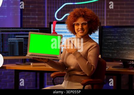 Lächelnde Entwicklerin präsentiert Laptop mit grünem Bildschirm, der den Software-Code im dunklen Büro anzeigt. Der Software-Code ist auf mehreren Computern sichtbar Stockfoto