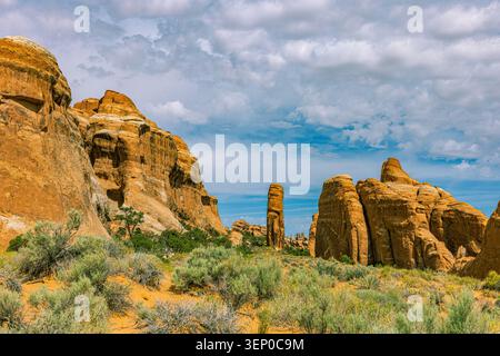 Im Arches National Park, Utah, befinden sich mächtige Sandsteinwände mit markanten hohen Felssäulen und Wüstensträucher unter bewölktem Himmel Stockfoto