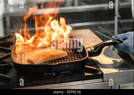 Gebratener Lachsfisch kocht in einer brennenden gusseisernen Pfanne. Flammen springen aus der Grillpfanne. Lachsfisch kocht intensiv. Grillvorgang bei hoher Hitze. Dampf steigt Stockfoto