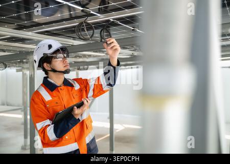 Junger Ingenieur in orangefarbener Uniform und weißem Helm inspiziert Solarpaneele und überprüft den Kabelanschluss unter Solarpaneelen, während er ein Tablet che hält Stockfoto