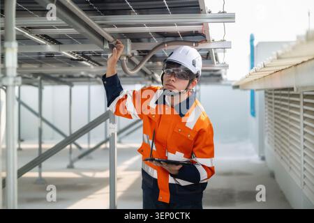 Ein junger Ingenieur inspiziert Solarpaneele an einer Metallkonstruktion. Trägt einen orangefarbenen Sicherheitsanzug und einen weißen Schutzhelm, hält ein Tablet und konzentriert sich auf die Ausrüstung Stockfoto