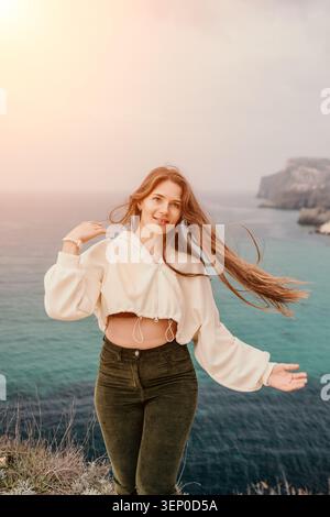 Frau Ozeanreise lächelnde junge Frau mit langen Haaren steht auf einer Klippe mit Blick auf das türkisfarbene Meer bei Sonnenuntergang Stockfoto