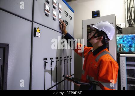 Asiatischer junger Elektroingenieur in orangefarbener, gut sichtbarer Jacke überprüft Bedienfelder. Der Mitarbeiter trägt einen weißen Schutzhelm und hält das Tablet. Bedienfelder f Stockfoto