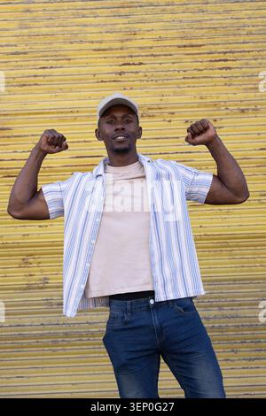 Erwachsener afroamerikanischer Mann, der mittig mit gelbem Verschluss steht, Arme biegt, Mütze und Jeans trägt Stockfoto