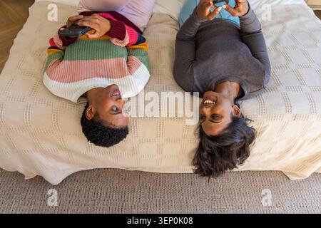 Verschiedene Freundinnen liegen auf dem Bett und halten Smartphones, in gestreiftem Pullover und grauem Oberteil lächelnd Stockfoto