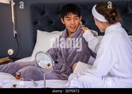 Verschiedene Paare in Bademänteln sitzen im Schlafzimmer, Frau, die Gesichtsrolle auf den Mann in der Nähe des Spiegels anwendet Stockfoto