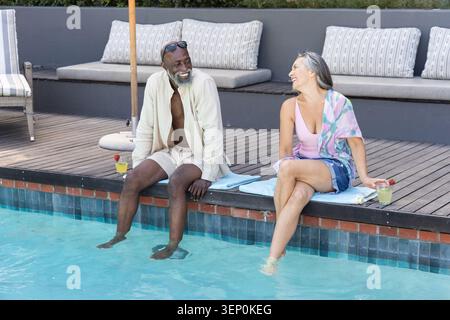 Verschiedene Paare sitzen auf gefliesten Poolrand auf hellblauen Handtüchern, tauchten Füße und halten Cocktails Stockfoto