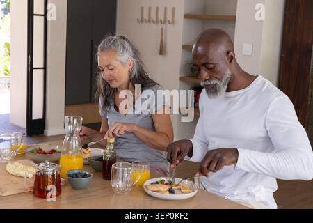 Senior Mann und Frau in grauen, weißen Hemden sitzen am Holztisch und essen Pfannkuchen mit Heidelbeeren Stockfoto