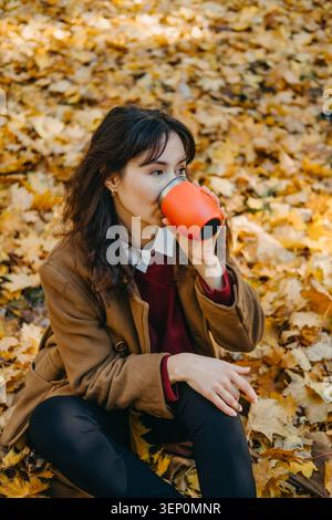 Die Frau sitzt auf einem mit Blättern bedeckten Boden und trinkt aus einem wiederverwendbaren Behälter. Wiederverwendbarer Thermobecher, Kaffeepause ohne Abfall, Becher für nachhaltige Reisen. Stockfoto