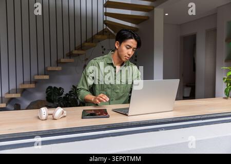 Asiatischer Mann, der hinter Holztheke zu Hause steht, mit silbernem Laptop, Tablet, Headset, in grünem Hemd Stockfoto