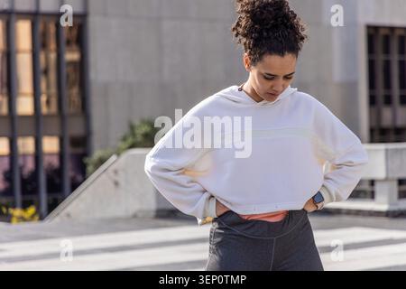 Frau in ihren Zwanzigern, die auf dem Dach stand, mit weißem Kapuzenpullover, Korallenoberteil, grauer Leggings Smartwatch Stockfoto