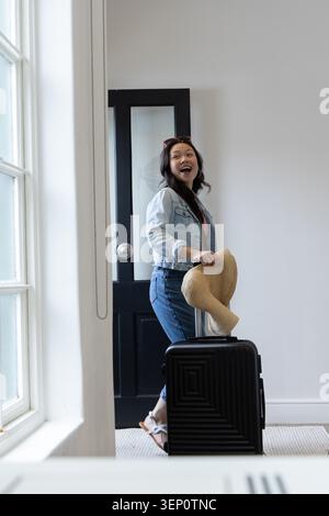 Asiatische Frau, die in hellem Eingangsbereich steht und einen Strohsonnenhut hält und einen schwarzen Koffer-Griff packt Stockfoto