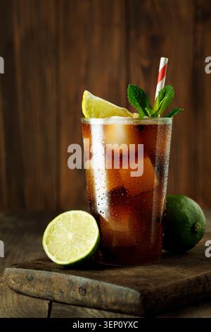 Erfrischender cuba Libre Cocktail mit Cola, Rum, Limette und Minze, kaltes Getränk mit Eiswürfeln auf rustikalem Holztisch Stockfoto