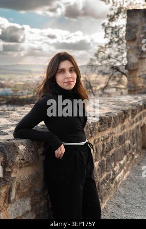 Vertikales Porträt einer jungen Frau, die sich an einer alten Steinmauer an einer historischen Stätte lehnt, mit sanfter Hintergrundbeleuchtung und bewölktem Himmel Stockfoto