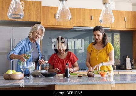 Verschiedene Freundinnen bereiten Mahlzeiten auf der Kücheninsel zu, hacken Gemüse und gießen Weißwein. Kochen, Gruppe, Zuhause, Zusammenarbeit, Stein, Granit, rostfrei Stockfoto