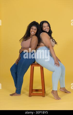 Verschiedene Frauen posieren Rücken an Rücken auf einem Holzhocker in gelbem Studio in kurzen Tanktops und Jeans. Porträt, Duo, Denim, barfuß, Hintergrund, minimalistisch, hochgradig Stockfoto