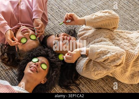 3 weibliche Freunde, die auf gewebtem Teppich liegen, tragen Pullover und Hoodie mit Gurkenscheiben, die die Uhr zeigen. Spa, Maske, Gesicht, Verwöhnung, Entspannung, Lounge, Freundschaft Stockfoto