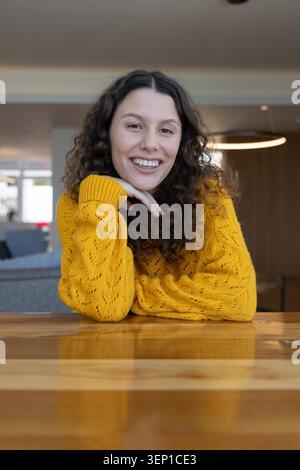 Frau Mitte 20 lehnte sich auf polierten Holztisch in der Lounge und lächelte in senfgelben Pullover. Wohnzimmer, gemütlich, Strickwaren, Anhänger, Innenausstattung, Lifestyle, Porträt Stockfoto