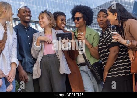 Verschiedene Freunde treffen sich im Glasbüro, elegant-lässig, sehen sich das Smartphone an und halten Kaffeetassen. Gruppe, Kollegen, Telefon, Outdoor, Urban, plaza, Gebäude Stockfoto