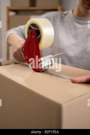 Nicht erkennbarer Arbeiter, der den Paketkarton mit Bandspender versiegelt. Schließen Sie die Hände an, indem Sie während der Verpackungsvorbereitung Verpackungsband anbringen, bestellen Sie für Shi Stockfoto