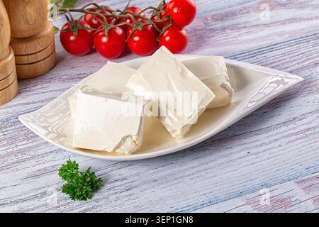 Weiße Fetakäswürfel auf Keramikschale mit roten Tomaten auf Weinrebe und grünen Kräutern auf rustikalem Holztisch Stockfoto