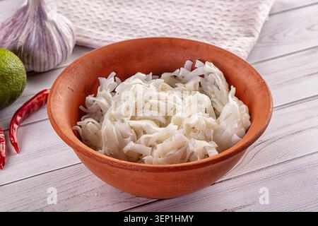 Nahaufnahme von rohen Reisnudeln in Terrakotta-Schüssel umgeben von Knoblauchzwiebeln Limette und roten Chilischoten auf weißem Holztisch mit sanfter natürlicher Beleuchtung Stockfoto