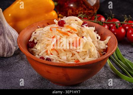 Traditioneller, fermentierter Kohlsalat gemischt mit Karotten und Preiselbeeren, serviert in einer rustikalen Schüssel aus Steingut, umgeben von frischem Gemüse, Knoblauch und tom Stockfoto