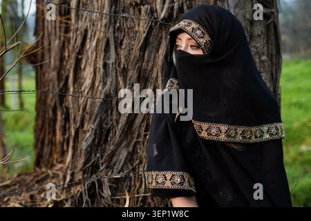 Muslimische Frau mit schwarzem Niqab mit Goldstickerei. Draußen stehen und zur Seite schauen. Mit Stacheldraht im Vordergrund und einem großen Baum darin Stockfoto
