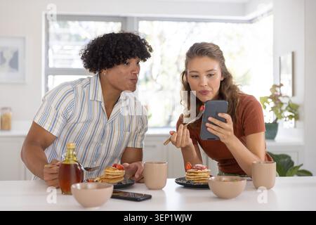 Pärchen, die an der Küchentheke sitzen und Pfannkuchen haben, Frau, die Smartphone und Gabel hält, Mann, der sich hineinlehnt Stockfoto