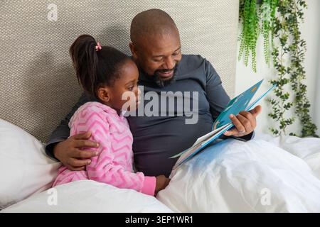 Afroamerikanischer Mann und Mädchen sitzen im Bett und lesen blaues Kinderbuch, Mädchen in rosa Gewand Stockfoto
