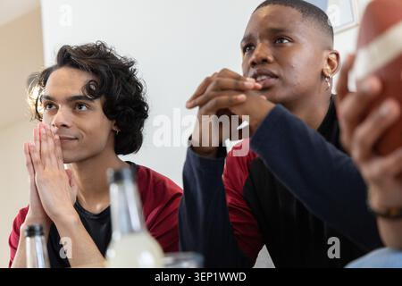 Verschiedene männliche Freunde, die zu Hause spielen, tragen kastanienbraune Hemden mit Flaschen Fußball auf dem Tisch Stockfoto