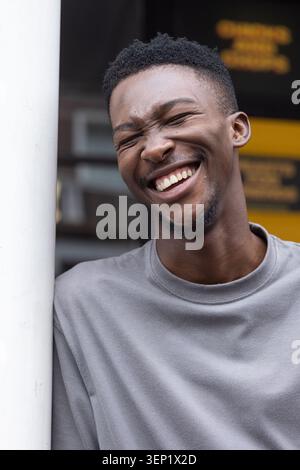Afrikanischer Mann lehnt sich an einen weißen Pfosten, lächelnd, Augen geschlossen am Ladenschild mit grauem Hemd Stockfoto