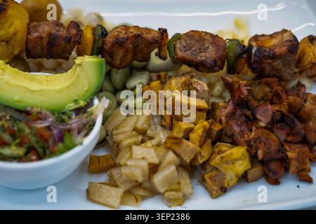Köstliche peruanische Anticuchos mit gegrilltem mariniertem Fleisch und Paprikaspießen, serviert mit geschnittener Avocado, Mais, Favabohnen und frittiertem Planta Stockfoto