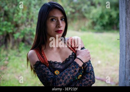 Junge Frau mit rotem Lippenstift und dunklem Haar, die aufmerksam in die Kamera blickte, ein besticktes Oberteil und Armstulpen trug und sich mit einem Arm umarmte Stockfoto