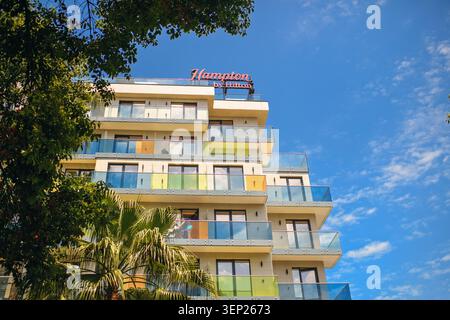 Batumi, Georgia - 09. Juni 2026: Blick auf die Fassade des Hampton by Hilton Hotelgebäudes mit farbenfrohen Glasbalkonen, eingerahmt von grünen Bäumen. Stockfoto