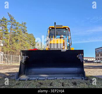 Gelber Baggerlader ist auf einer Baustelle geparkt. Der Frontlader zeigt nach vorn mit einem klaren blauen Himmel über dem Kopf. Bäume und Gebäude sind in t zu sehen Stockfoto