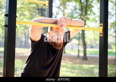 Besinnlicher Mann mit Brille im Park. Ein Mann in schwarzem Hemd hält an, um unter Bäumen nachzudenken. Weißer bärtiger Mann mit Gläsern, die nachdenklich an der Bar liegen, umgeben von Parkbäumen Stockfoto
