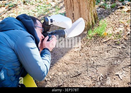 Der Naturfotograf kniet auf dem Waldboden, um Nahaufnahmen mit der Kamera im Frühjahr im Waldlicht aufzunehmen. Nützlich für Outdoor-Marketing, Hobbywerbung, Bildung und Lifestyle-Konzepte Stockfoto