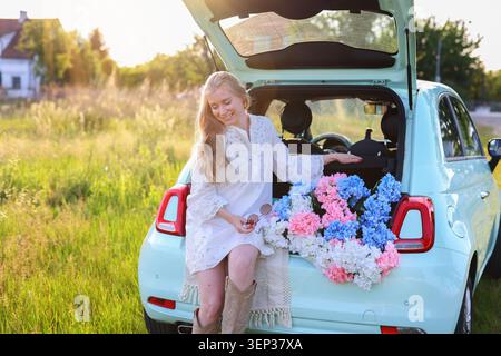 Lächelnde junge Frau, die zur goldenen Stunde im Kofferraum eines pastellblauen Autos mit Hortensien sitzt und einen Sommerabend auf dem Land genießt Stockfoto