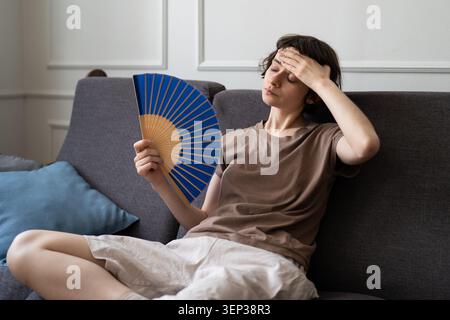 Junge Frau, die auf dem Sofa liegt und mit einem Handpapierfächer winkt, unter Sommerhitze leidet, Kopfschmerzen empfindet. Stockfoto
