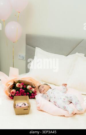 Das Neugeborene schläft friedlich auf einem rosafarbenen Kissen mit einem Blumenstrauß und Ballons in der Nähe, auf einem weißen Bett in einer gemütlichen Innenumgebung Stockfoto