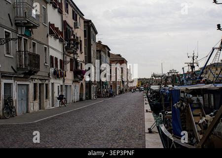 Eine enge Straße am Wasser in Chioggia, Italien, gesäumt von Fischerbooten, Wohnhäusern, Fahrrädern und alltäglichem Hafenleben. Stockfoto