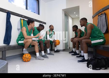 Verschiedene männliche Teamkollegen in grünen Trikots sitzen auf Bänken in der Umkleidekabine und halten Basketball Stockfoto