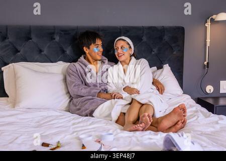 Verschiedene Paare sitzen auf dem Bett und halten Hände, tragen Bademäntel, blaue Flecken, zeigen Hautpflegegläser Stockfoto