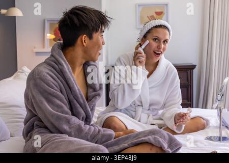 Verschiedene Paare, die in Bademänteln auf dem Bett sitzen, eine Frau, die ein tragbares Gesichtsgerät und eine Brille hält Stockfoto
