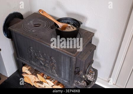 Traditioneller Holzofen mit Eisentopf mit Haferbrei, Holzlöffel und gestapeltem Brennholz im Innenbereich Stockfoto
